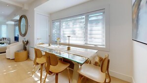 Een moderne eetruimte met een glazen tafel, houten stoelen en een groot raam met jaloezieën.