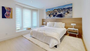 Een slaapkamer met een groot bed, wit beddengoed, een houten hoofdbord, een schilderij boven het bed en een raam met jaloezieën.