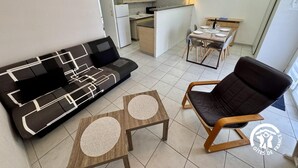 En kompakt opholdsstue med sofa, stol, lille bord, tekøkken og spiseplads.