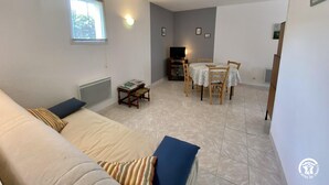 A living room with a sofa, a small table with books, a TV stand, and a dining area with a table and chairs. - Camplong l'Alaric - CAMPLONG D'AUDE (CAMPLONG D'AUDE)