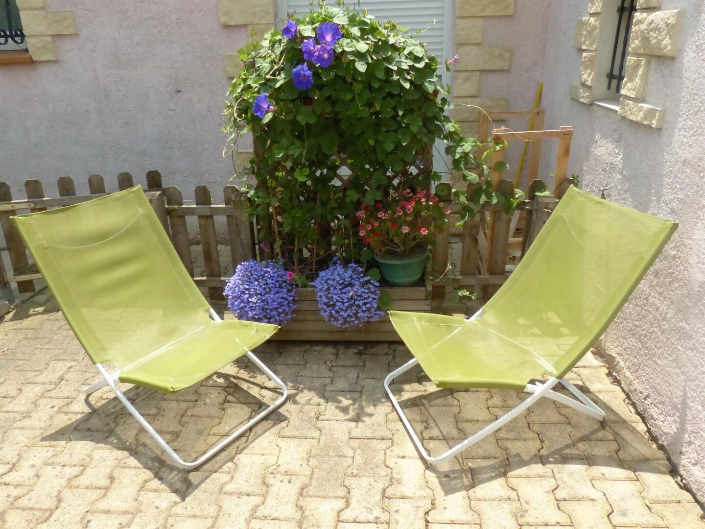 Deux chaises de jardin vertes sont placées sur une terrasse dallée, avec une clôture en bois en arrière-plan.