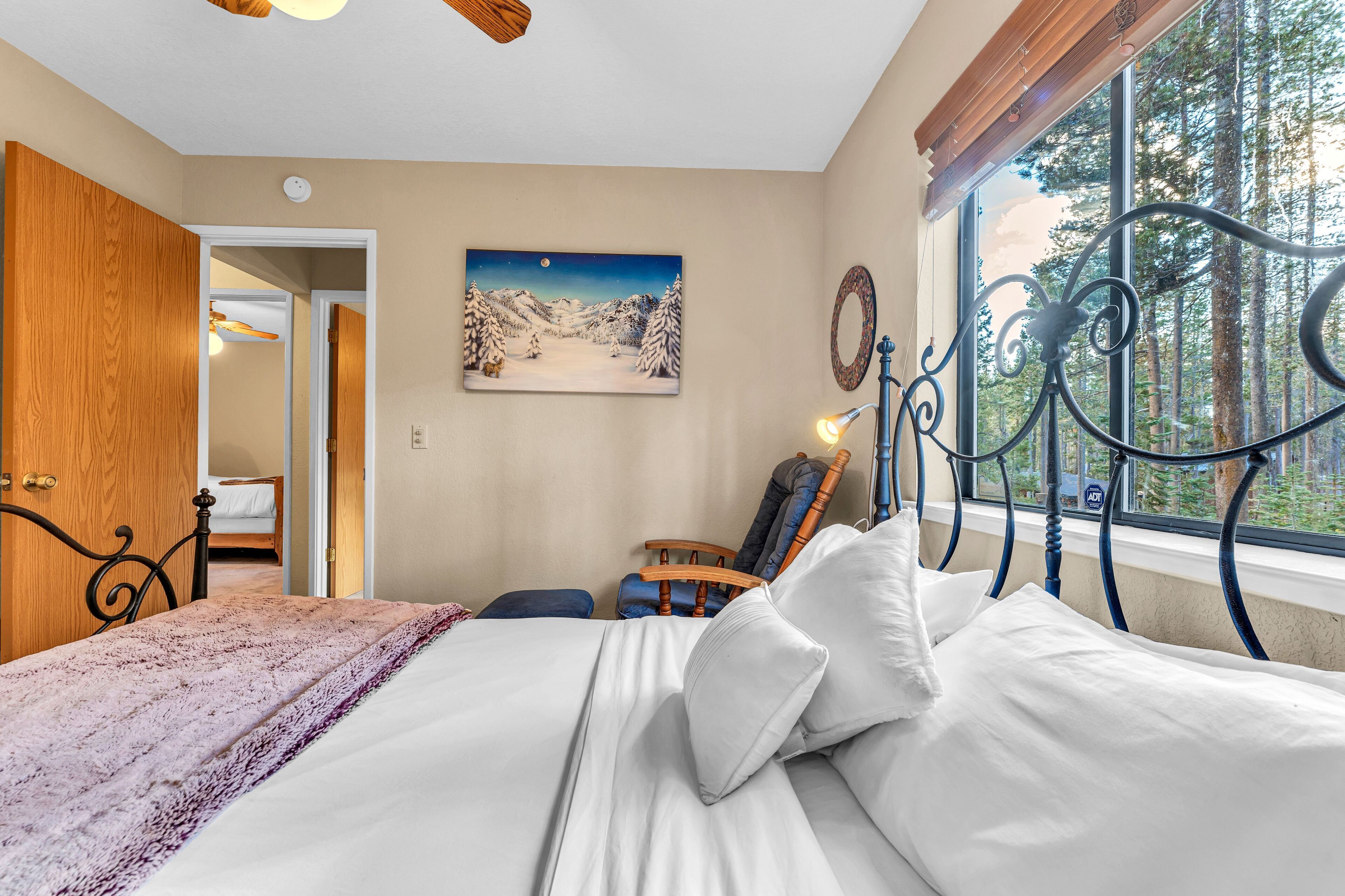 A bedroom with a bed, a wooden door, a painting, a window with a view of trees, and a metal headboard.