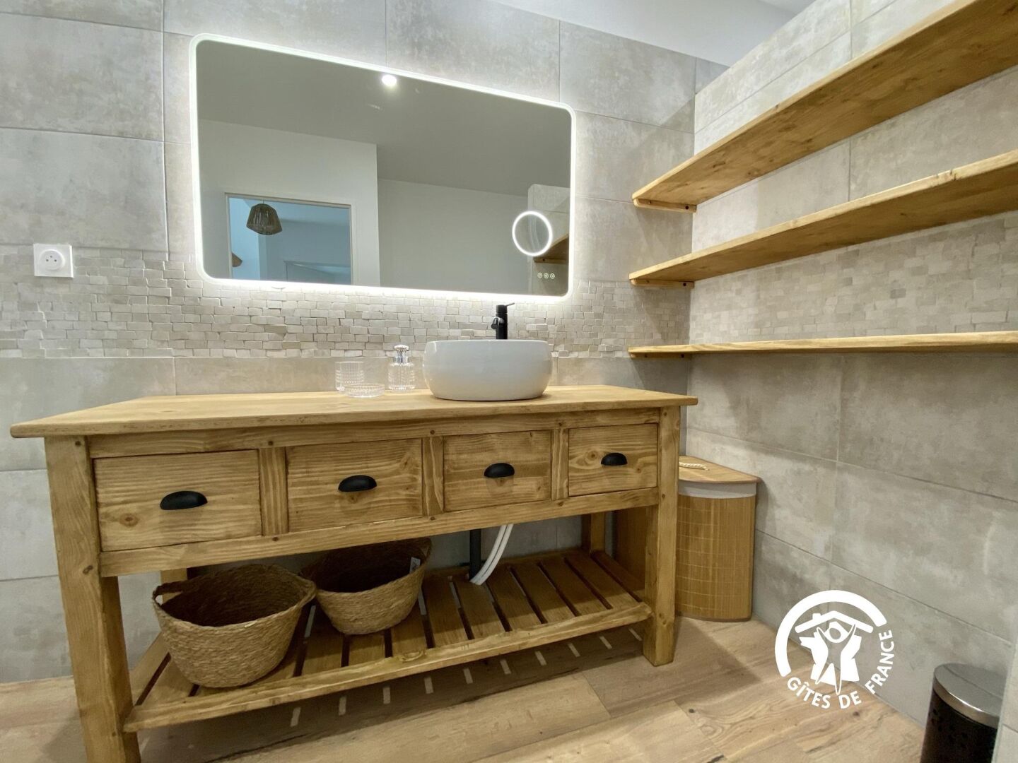 A modern bathroom with a wooden vanity, a white sink, and a mirror with a light. There are wooden shelves above the vanity and a trash bin to the side.