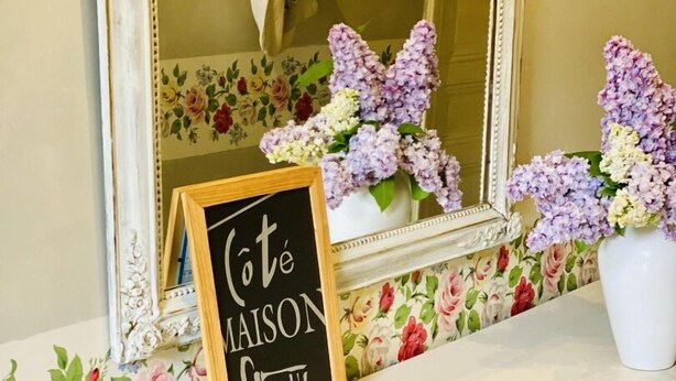 Uma área de recepção de hotel com papel de parede floral, um espelho, uma lousa e um vaso de flores, além de revistas de viagem no balcão.