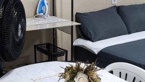 Una habitación con una mesa blanca, un ventilador, una tabla de planchar, una cama con ropa de cama gris y una silla blanca.