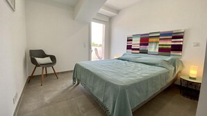 A bedroom with a bed, a chair, a colorful artwork on the wall, and a window. - L'ancienne Cave - ROQUEFORT DES CORBIERES (ROQUEFORT DES CORBIERES)