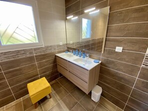 Une salle de bain moderne avec un grand miroir, un lavabo et des toilettes. Une fenêtre laisse entrer la lumière naturelle et il y a un tabouret jaune.