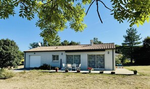 Une maison de plain-pied avec un toit en tuiles, un garage et une terrasse aménagée avec du mobilier d’extérieur.