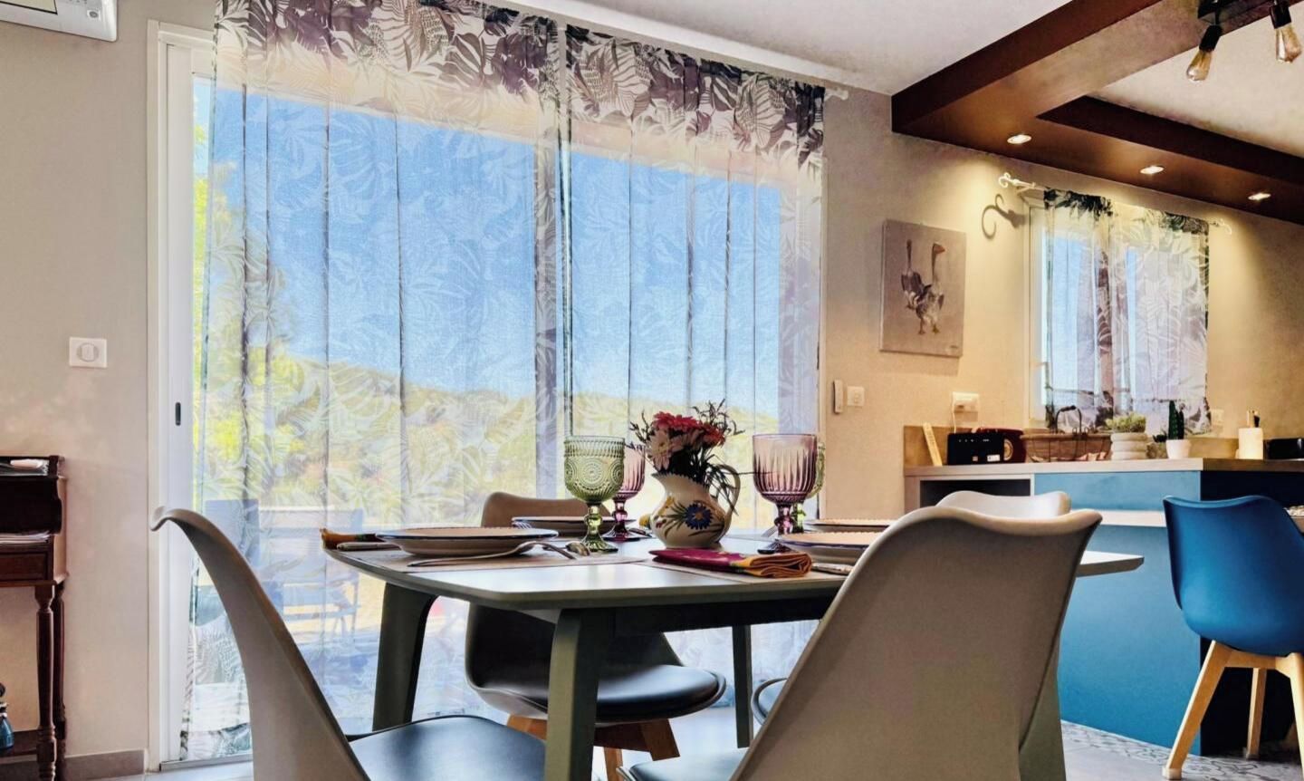 A dining area with a table set for two, a vase with flowers, and a view through large windows.