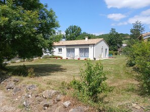 Une maison de plain-pied avec un toit en tuiles, un porche à l’avant avec une table et des chaises, et un petit jardin agrémenté de rochers et de plantes.