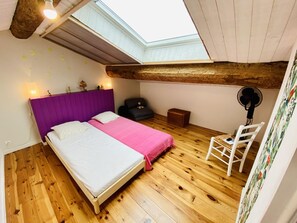 A bedroom with a sloped ceiling, wooden flooring, a bed with a purple headboard, a small table, a chair, and a fan. - Terre et Mer - ROQUEFORT DES CORBIERES (ROQUEFORT DES CORBIERES)