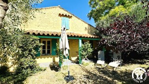 A rustic building with a tiled roof, green shutters, and a garden area with a bench and a chair. - Le Mas des Oliviers - AZILLE (AZILLE)