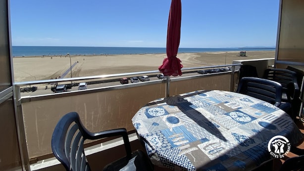 Un balcon avec une table, des chaises et un parasol rouge, donnant sur une plage.