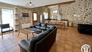 Un salon avec un canapé en cuir noir, un coin repas avec une cuisine en arrière-plan, des murs en pierre et une grande fenêtre.