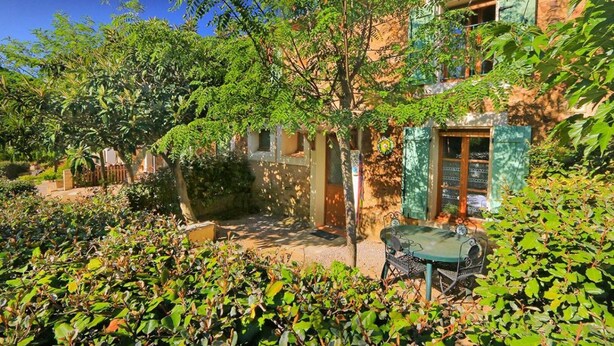 Un cortile con un edificio rustico, circondato da vegetazione lussureggiante e un tavolo in metallo.