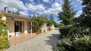 Une maison avec une allée en gravier, un patio couvert avec une table et des chaises, et un grand arbre en arrière-plan.