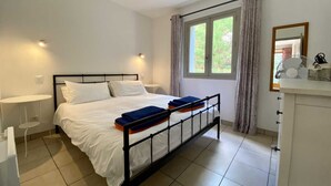 Une chambre d’hôtel comprenant un lit, une table de chevet, une fenêtre avec des rideaux, un miroir et un petit bureau avec un ventilateur.