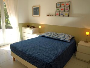 A bedroom with a bed, bedside tables, a nightstand, a wall-mounted shelf with decorative items, and a window with curtains. - Vista Alegre - FITOU (FITOU)