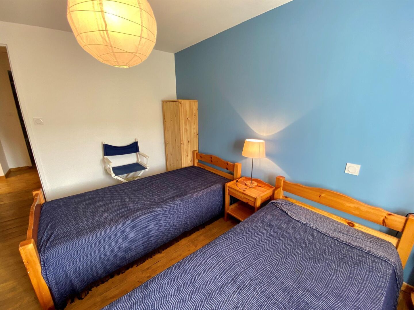 Ein Zimmer mit zwei Holzbettgestellen mit blauer Bettwäsche, einem Nachttisch aus Holz mit einer Lampe, einer blauen Wand und einem Holzschrank.