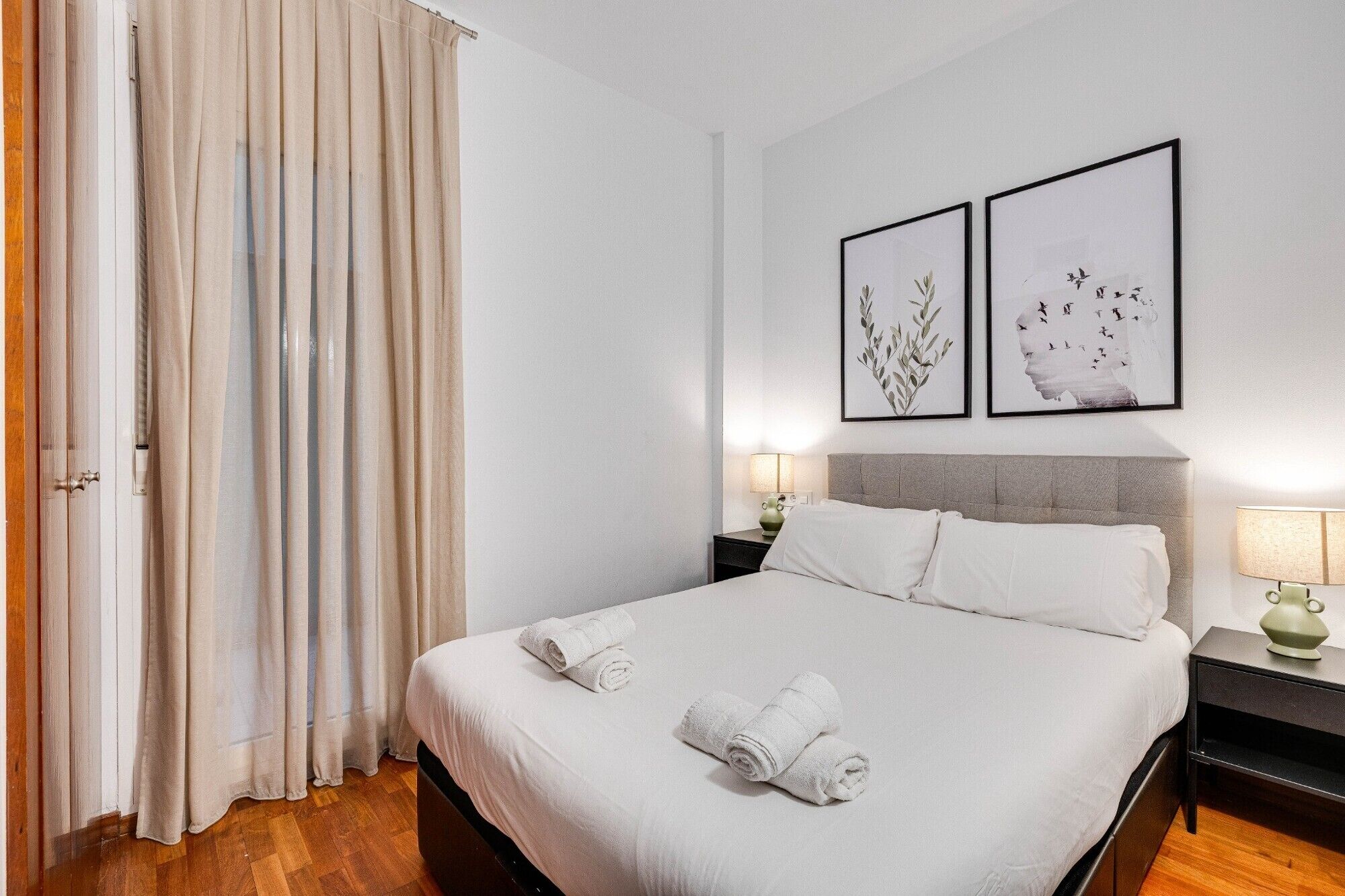 Un lit bien fait, avec du linge de lit blanc et des serviettes pliées, une tête de lit grise et deux tables de chevet avec des lampes.