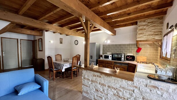 A cozy interior with a stone island counter, wooden ceiling beams, a dining area with chairs, and a kitchen with a microwave and stove. - La Bouriette - MOUSSOULENS (MOUSSOULENS)