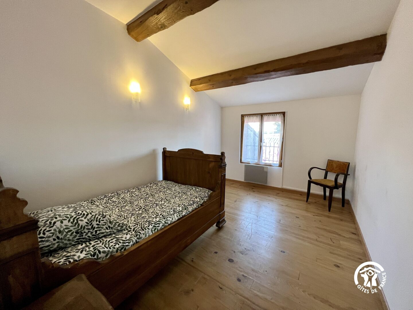A small, single-bed room with a wooden floor, a window with curtains, and a chair.