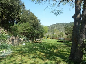A grassy outdoor area with a swing set and a table, surrounded by trees and a stone wall. - L'Oustalou - SAINT MARTIN DES PUITS (SAINT MARTIN DES PUITS)