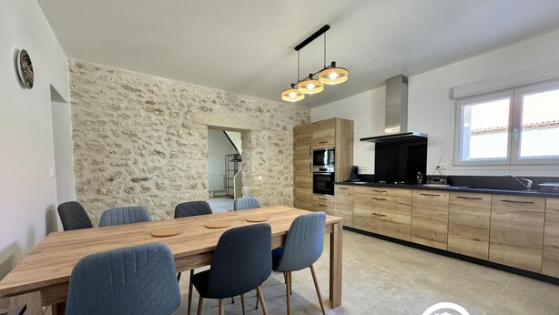 A modern kitchen with a stone wall, wooden cabinets, and a dining area with a long table and chairs. - Etoiles des Salins - Gîte 2 (PORT LA NOUVELLE)