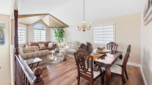 Una sala de estar amplia con techo alto, un comedor con mesa y sillas de madera, y una escalera con barandilla de madera.