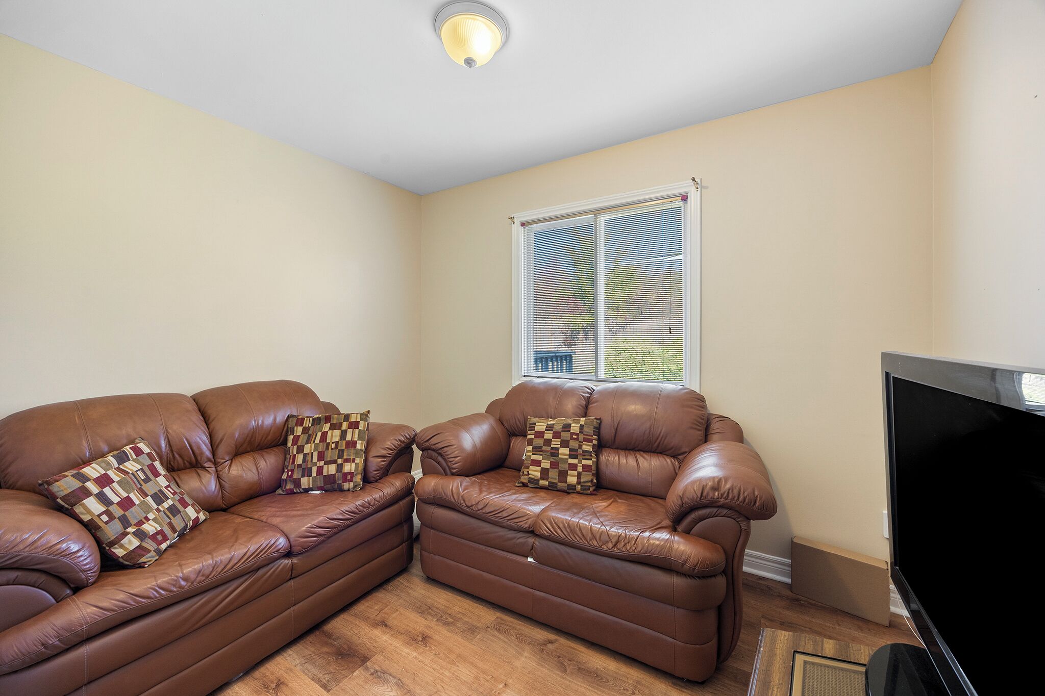 A living room with two brown leather sofas, patterned throw pillows, a large flat-screen TV, and a window with blinds.