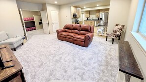 Ein Wohnzimmer mit einem braunen Ledersofa, einem weißen Sessel und einem Holztisch. Im Hintergrund ist eine Küche mit Holzschränken und einem Edelstahl-Kühlschrank zu sehen.