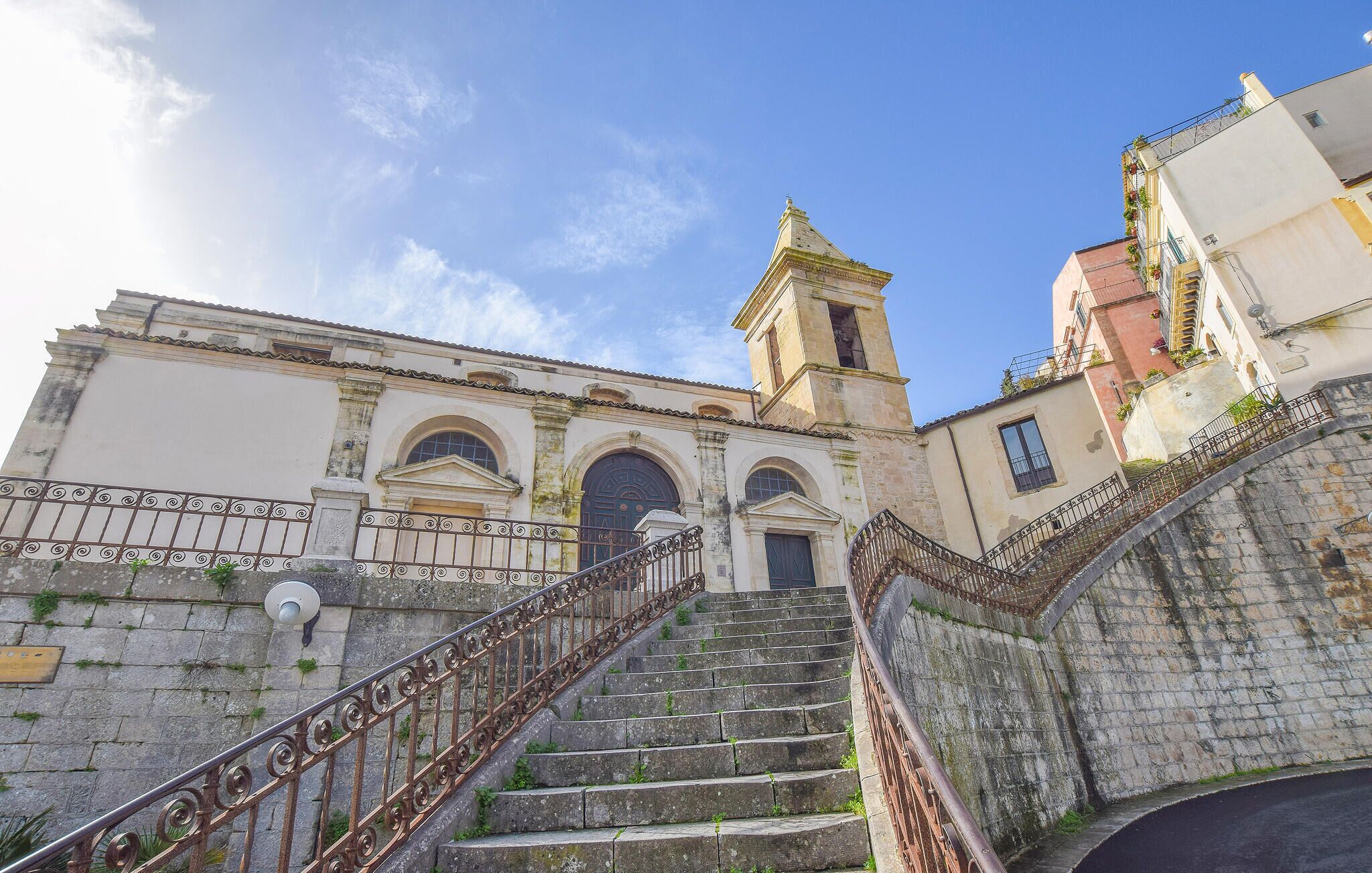 Un edificio histórico con un campanario, una escalera que conduce a la entrada y un muro de piedra con barandilla de metal.