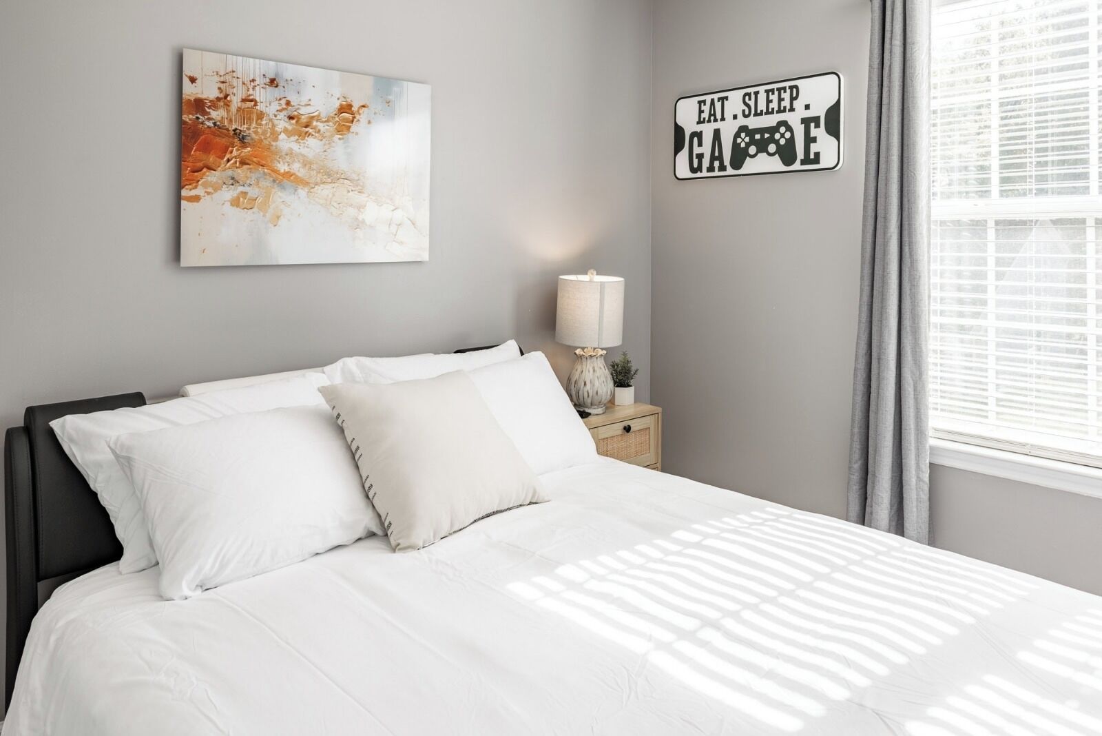 Een slaapkamer met een bed, een nachtkastje met een lamp, een kunstwerk aan de muur en een bordje op de muur.