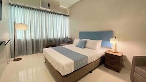 A modern hotel room with a large bed, a bedside table with a lamp, a chair, and a window with curtains. - UPad Hotel (Manila)