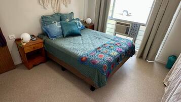 A bedroom with a bed, a nightstand, a ceiling fan, a window with curtains, and a wall-mounted macrame decoration.