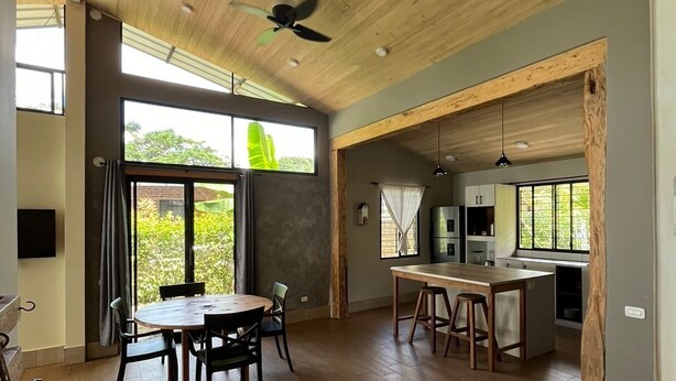 Una cocina amplia con piso de madera, un comedor con mesa redonda y ventanas grandes que ofrecen vistas a la vegetación.