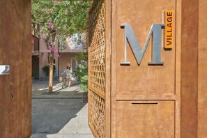 A wooden gate with a large 'M' and 'VILLAGE' sign, leading to a courtyard with a person walking. - Express by M Village Nam Ky Khoi Nghia, a brand of Modern Village Lifestyle (Ho Chi Minh City)