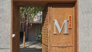 A wooden door with a large 'M' and 'VILLAGE' sign, indicating a hotel entrance. - Express by M Village Nam Ky Khoi Nghia, a brand of Modern Village Lifestyle (Ho Chi Minh City)