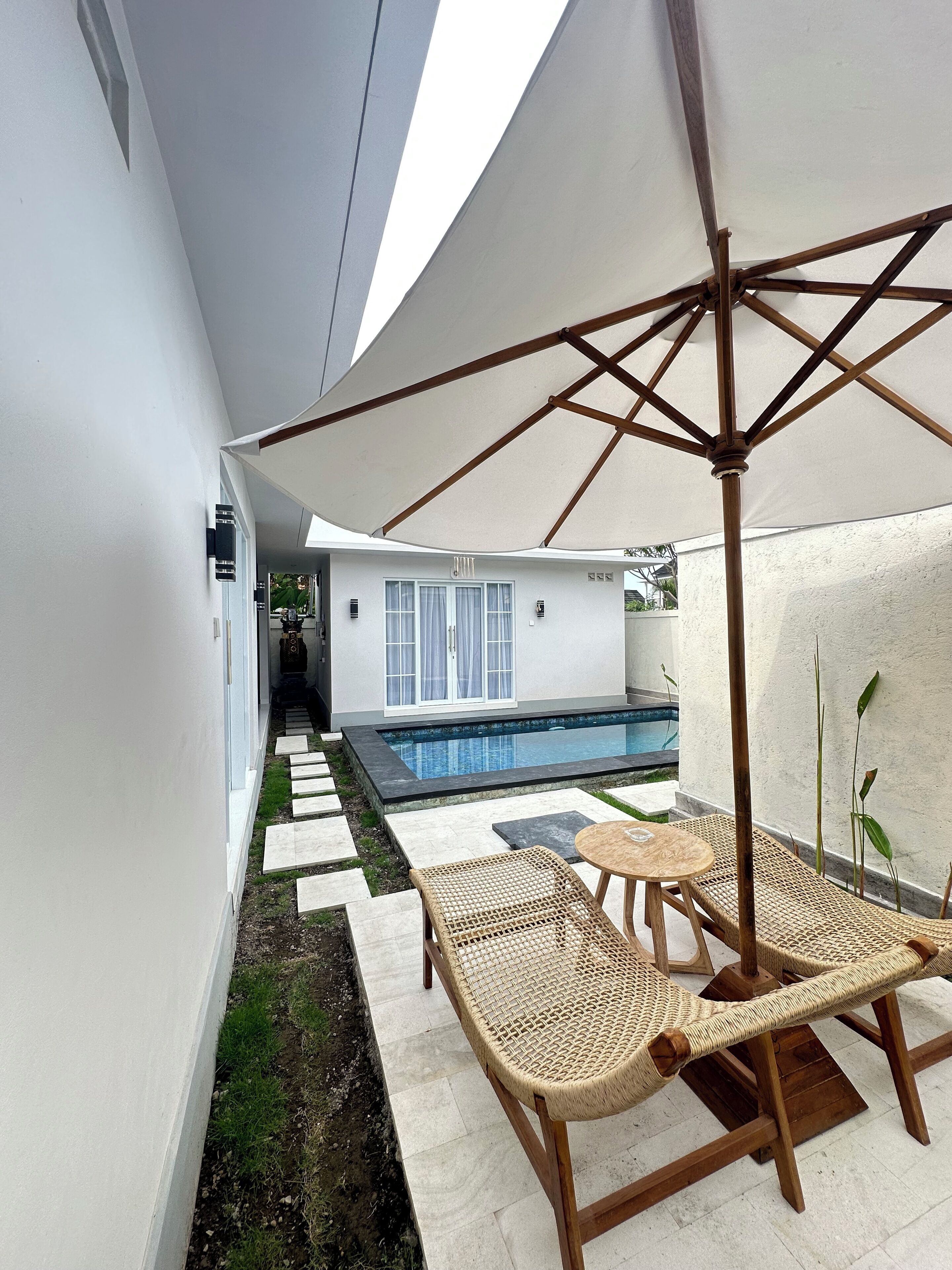 Uma área à beira da piscina com parede branca, piscina e um espaço coberto com mesa e cadeiras.