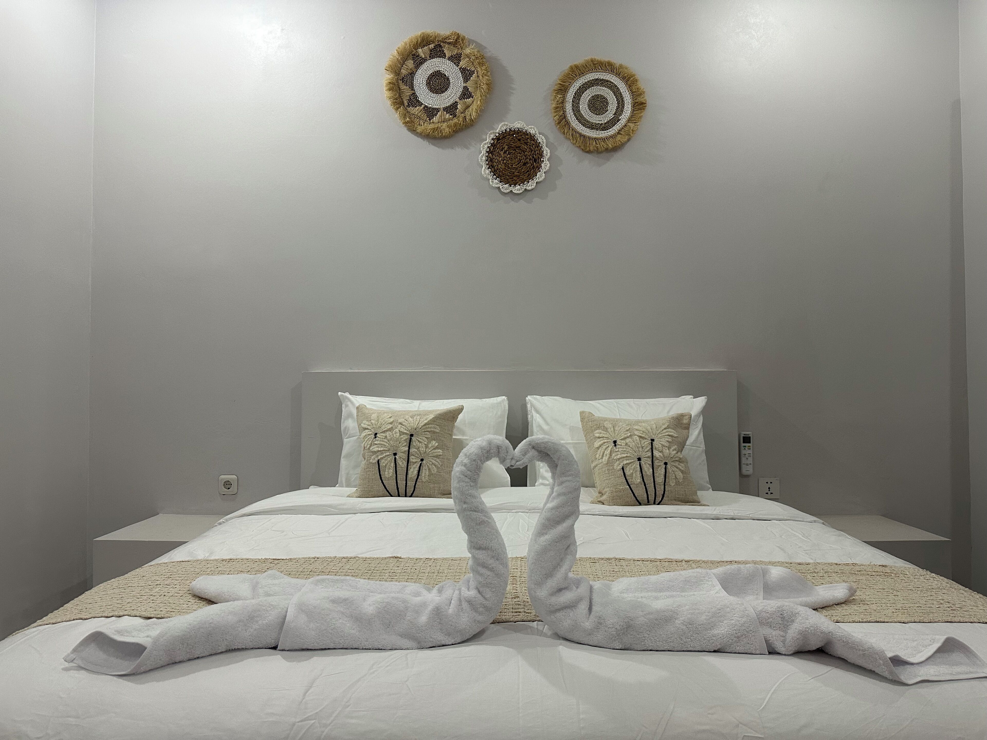 Uma cama bem arrumada com duas toalhas dispostas em formato de coração, uma cabeceira e três objetos decorativos fixados na parede.
