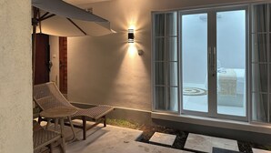 Un espace extérieur accueillant avec des fauteuils en osier et une table, une grande fenêtre avec des rideaux et un luminaire fixé au mur.