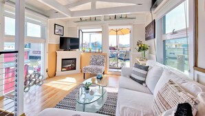Un salon spacieux avec un canapé blanc, une table basse en verre et une cheminée. Un téléviseur est fixé au mur, et une grande fenêtre offre une vue sur la ville.