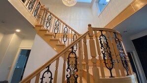 Un magnifique escalier, orné de rampes noires travaillées et de balustres en bois, mène à un plafond haut percé d’une lucarne et surmonté d’un lustre décoratif.