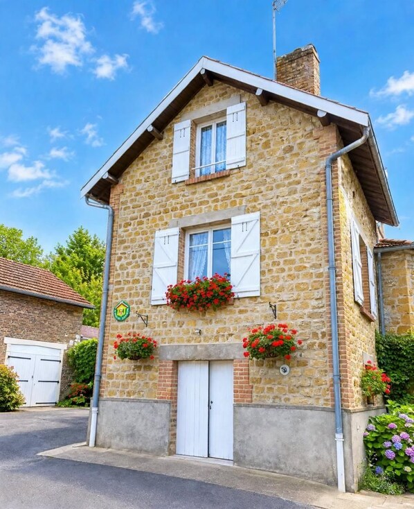 Una casa de piedra con una puerta blanca, dos ventanas con contraventanas y macetas con flores en la fachada.