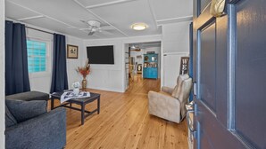 A living room with a sofa, armchair, coffee table, and a TV. There is a ceiling fan, a window with curtains, and a door with a decorative handle. - Historic Downtown New Bern Retreat - Pet Friendly (New Bern)