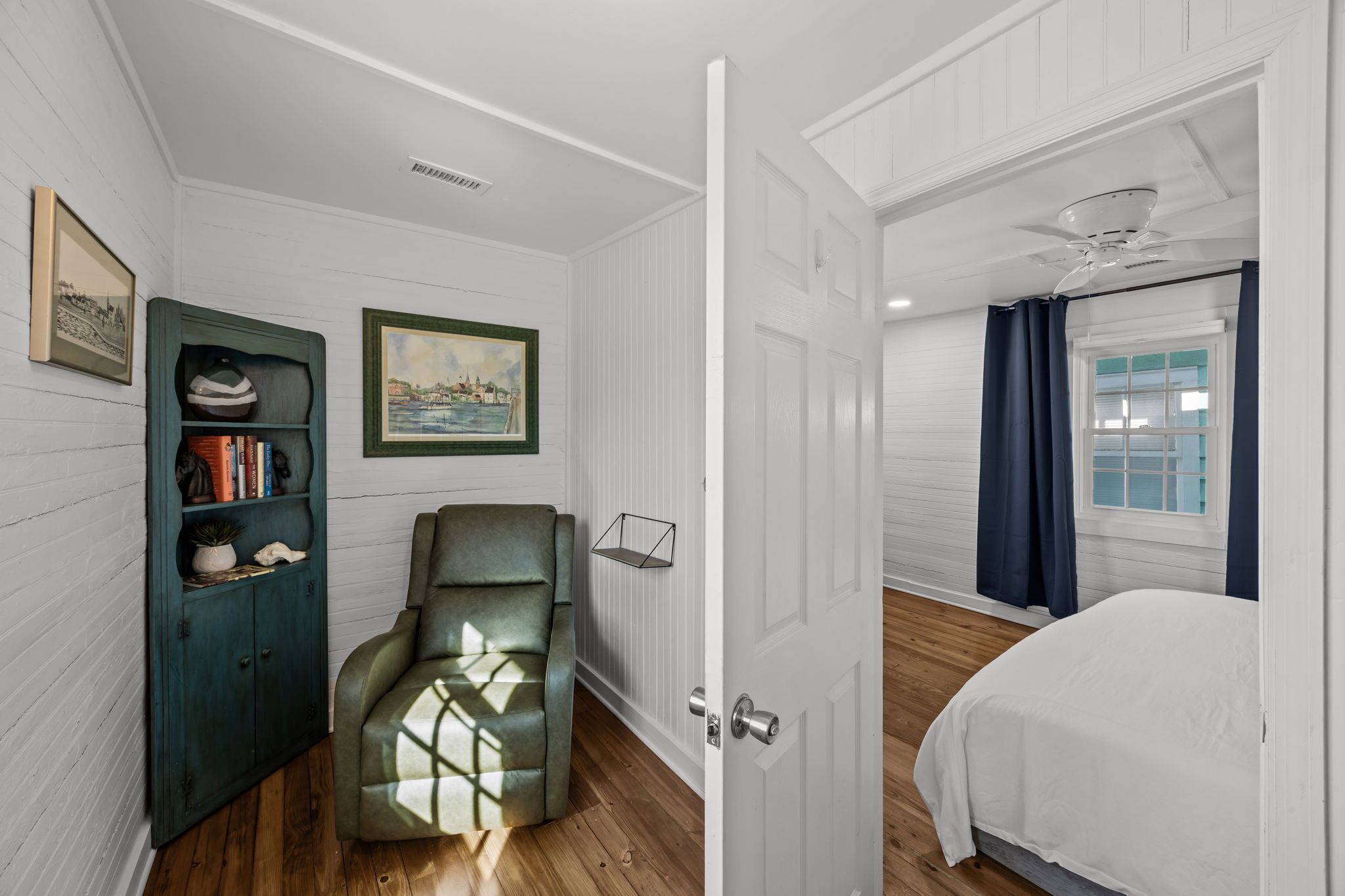 A bedroom with a bed, a green armchair, a bookshelf, a framed picture, and a window with curtains.