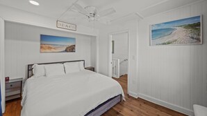 A bedroom with a bed, a ceiling fan, a painting of a beach, and a door leading to another room. - Historic Downtown New Bern Retreat - Pet Friendly (New Bern)