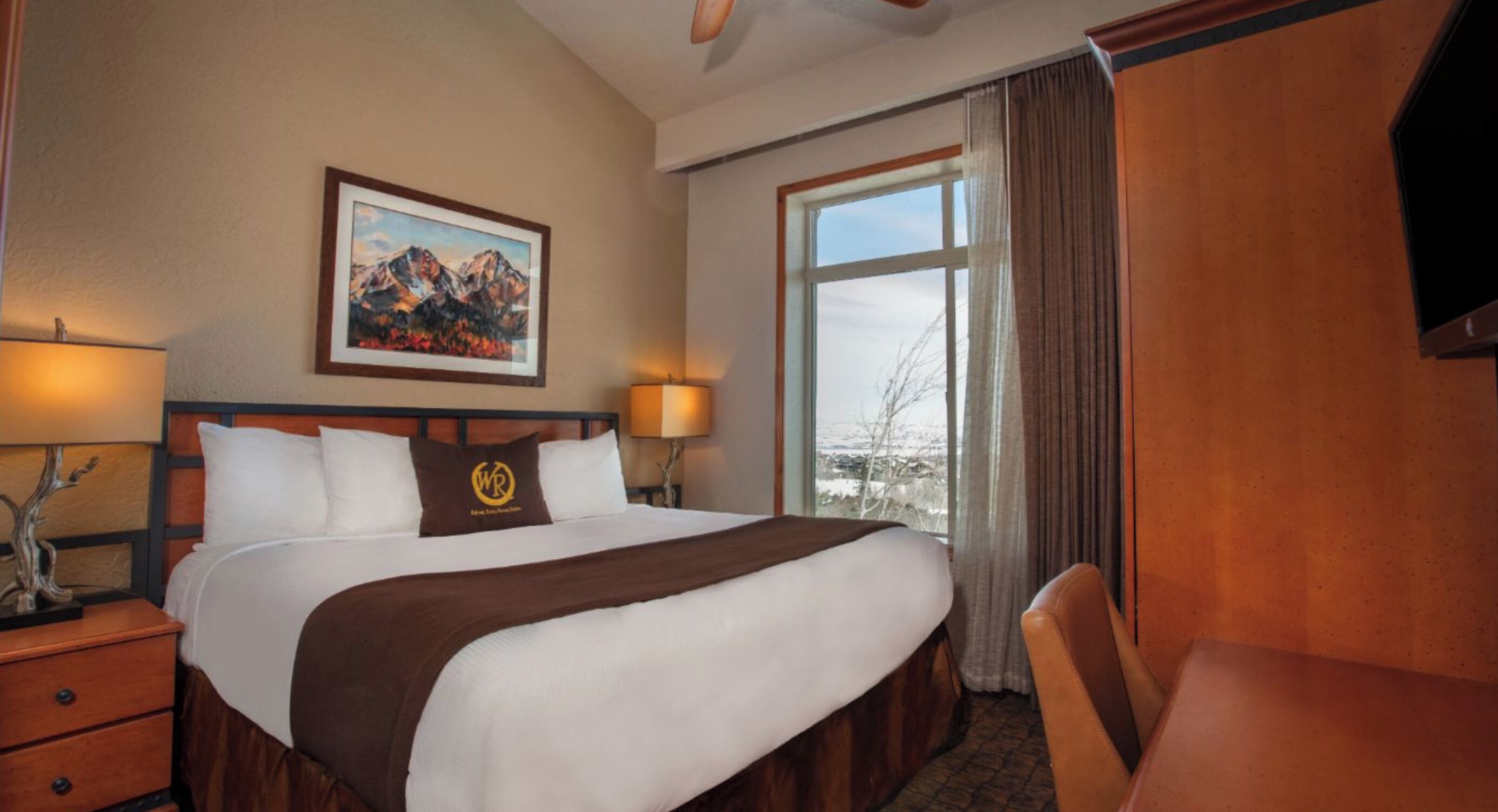 Habitación de hotel con una cama grande, dos lámparas de noche, un cuadro de montañas, una ventana con cortinas y un armario de madera.