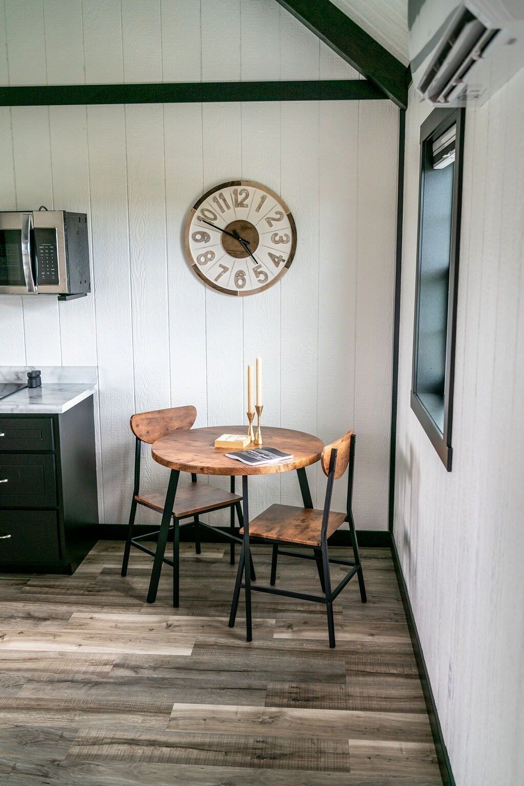 Una pequeña cocina con un reloj de madera, un microondas y una mesa redonda con dos sillas.