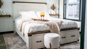 Ein ordentlich bezogenes Bett mit einer weißen und beige farbenen Decke, ein Nachttisch mit zwei Gläsern Orangensaft und ein kleiner Hocker.